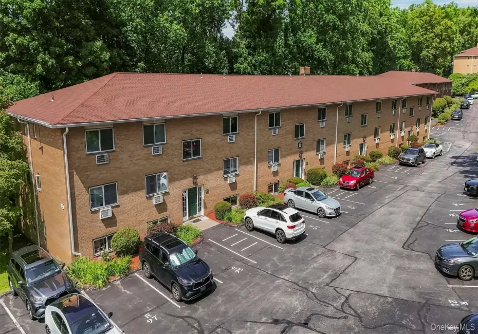 Brick facade building featuring multiple residential units, a red shingle roof, and a dedicated asphalt parking area with marked spaces Brick facade building featuring multiple residential units, a red shingle roof, and a dedicated asphalt parking area with marked spaces