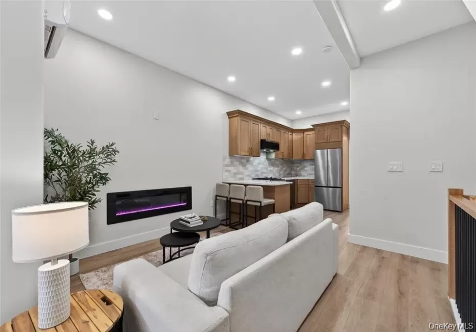 Open-concept living area featuring an electric fireplace, wood-finish flooring, recessed lighting, and a kitchen with wood cabinetry and stainless steel appliances Open-concept living area featuring an electric fireplace, wood-finish flooring, recessed lighting, and a kitchen with wood cabinetry and stainless steel appliances