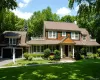 Expansive lawn with mature landscaping, a two-car garage with arched doors, a covered entry with white columns, and wood shake siding with white trim Expansive lawn with mature landscaping, a two-car garage with arched doors, a covered entry with white columns, and wood shake siding with white trim