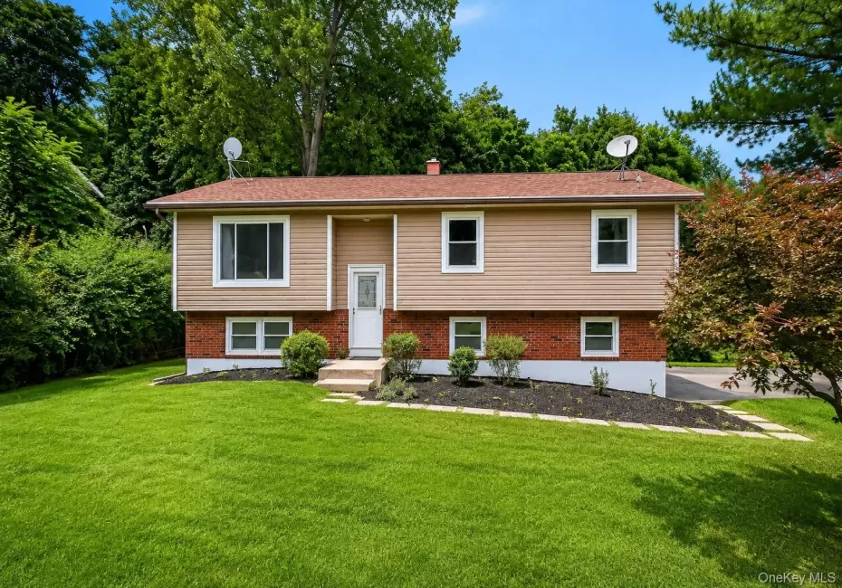 Split-level home featuring a brick and vinyl exterior with multiple windows, a centered entrance with concrete steps, and a professionally landscaped front yard with mature trees, manicured lawn, and established plantings along the foundation Split-level home featuring a brick and vinyl exterior with multiple windows, a centered entrance with concrete steps, and a professionally landscaped front yard with mature trees, manicured lawn, and established plantings along the foundation