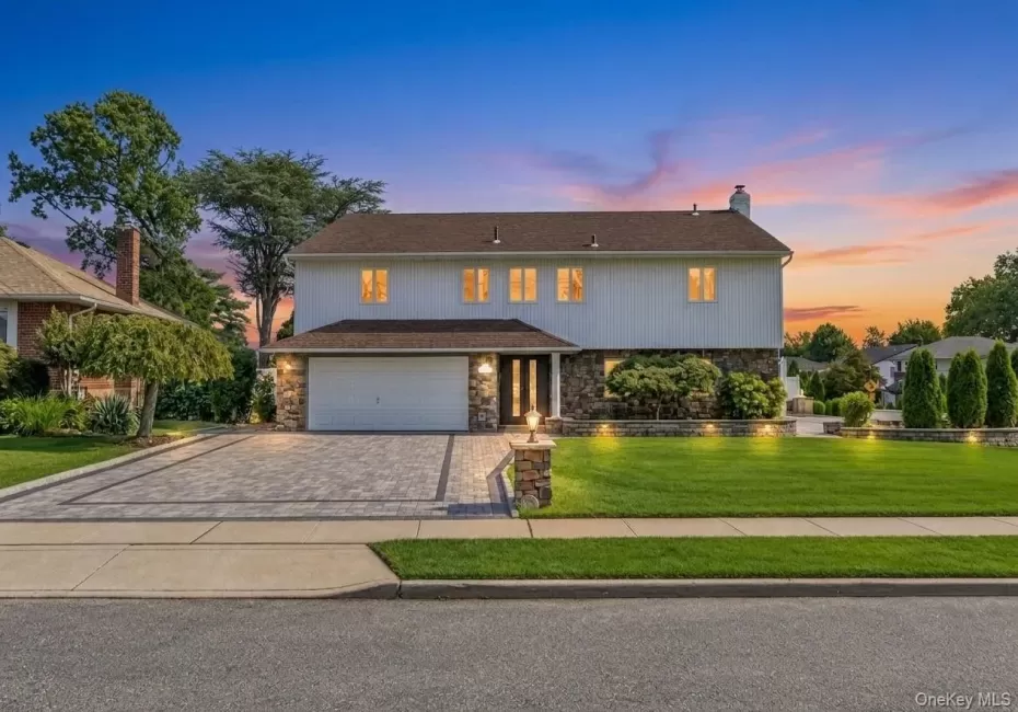 Two-story residence featuring a paved driveway, stone facade accents, an integrated garage, a manicured lawn, and exterior lighting Two-story residence featuring a paved driveway, stone facade accents, an integrated garage, a manicured lawn, and exterior lighting