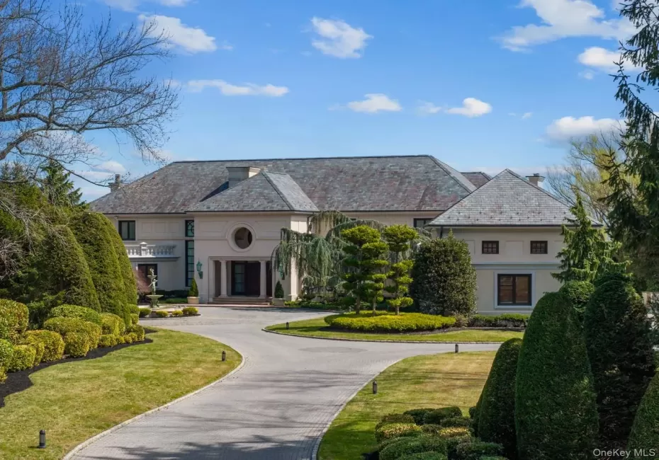 Grand estate featuring a circular driveway, manicured landscaping, a slate-style roof, a portico entrance, and an upper-level balcony with balustrade Grand estate featuring a circular driveway, manicured landscaping, a slate-style roof, a portico entrance, and an upper-level balcony with balustrade