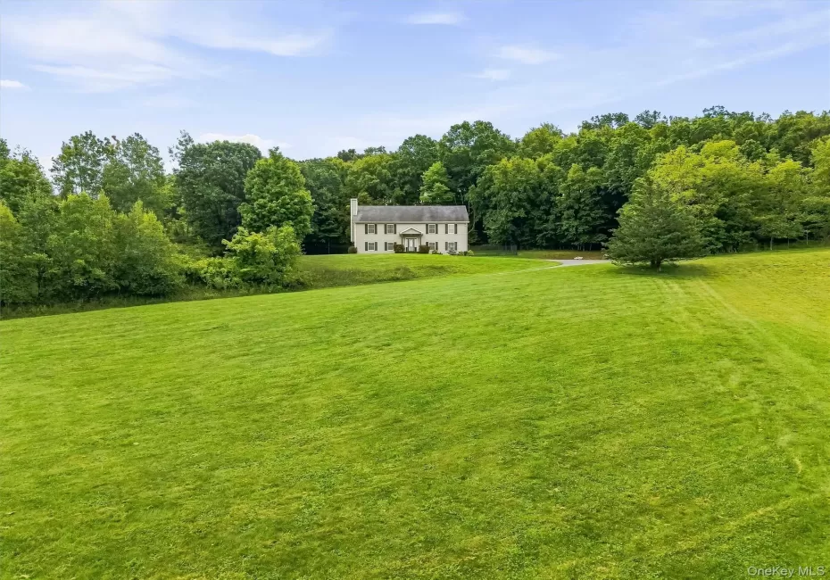 Expansive green lawn surrounds the property, featuring a traditional style home with a light-colored facade and dark shutters, set against a backdrop of mature trees Expansive green lawn surrounds the property, featuring a traditional style home with a light-colored facade and dark shutters, set against a backdrop of mature trees