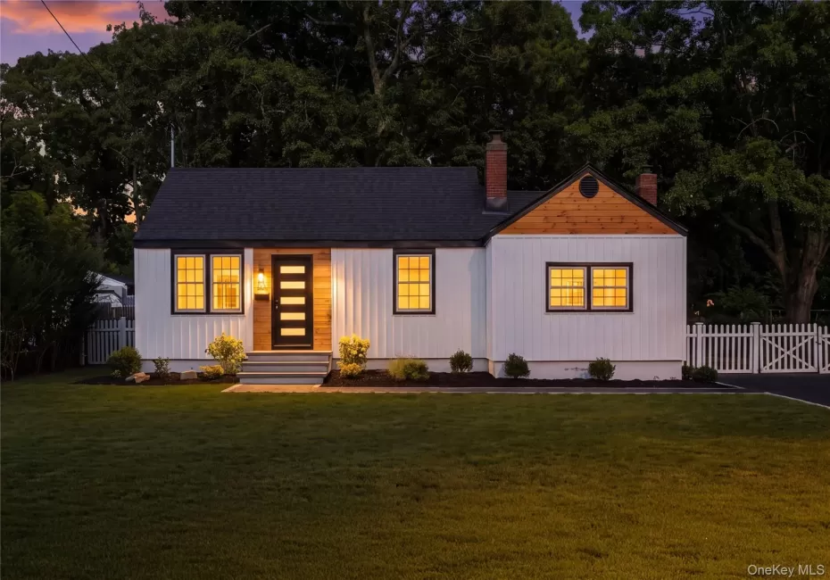 Front exterior featuring white vertical siding, dark framed windows, a dark shingle roof, a wood-paneled entry accent, and a white picket fence Front exterior featuring white vertical siding, dark framed windows, a dark shingle roof, a wood-paneled entry accent, and a white picket fence