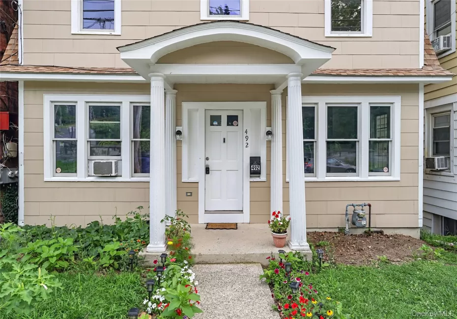 Columned portico entrance with a concrete pathway, flanked by garden beds, and light-toned siding Columned portico entrance with a concrete pathway, flanked by garden beds, and light-toned siding