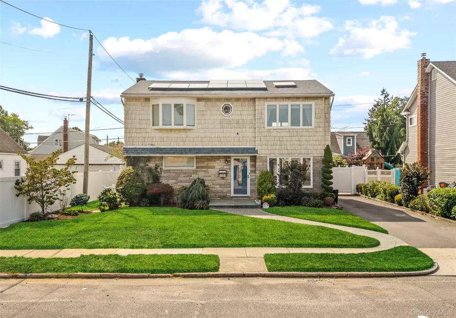 Two-story residence featuring a shingled exterior and stone facade, solar panels, a paved driveway, and a front lawn with a concrete pathway Two-story residence featuring a shingled exterior and stone facade, solar panels, a paved driveway, and a front lawn with a concrete pathway