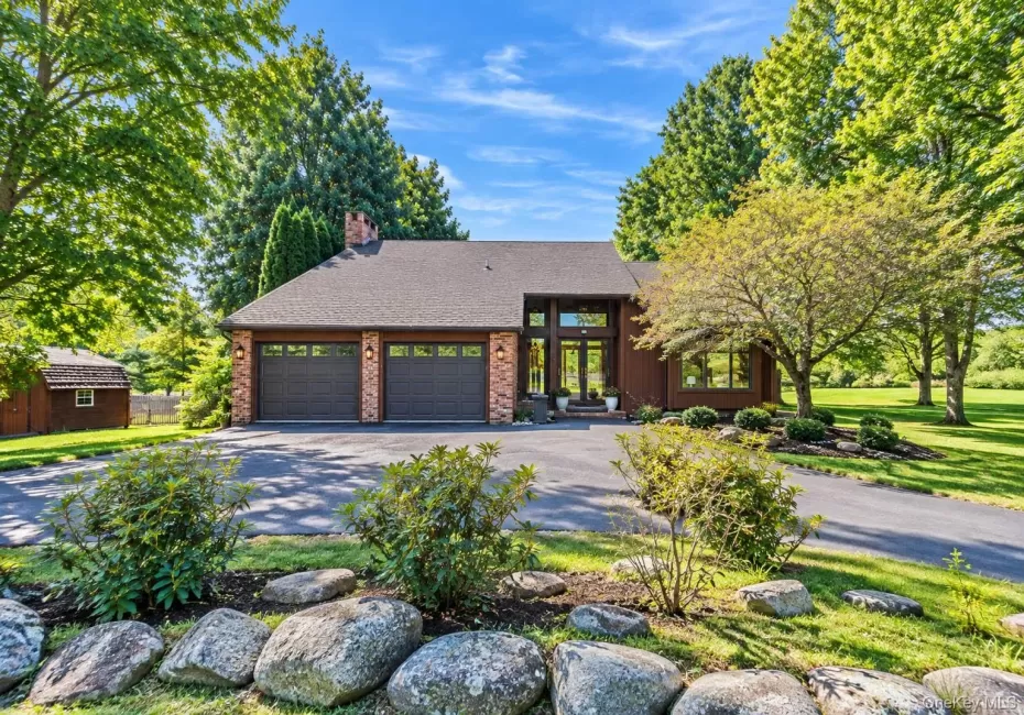 Property exterior featuring a multi-car garage, brick facade accents, a prominent front entrance with transom windows, and mature trees Property exterior featuring a multi-car garage, brick facade accents, a prominent front entrance with transom windows, and mature trees