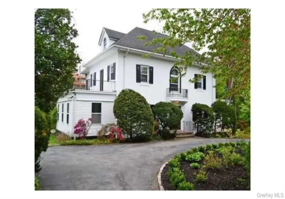 White exterior with black shutters, dormer window, second-story balcony with metal railing, and a paved driveway White exterior with black shutters, dormer window, second-story balcony with metal railing, and a paved driveway
