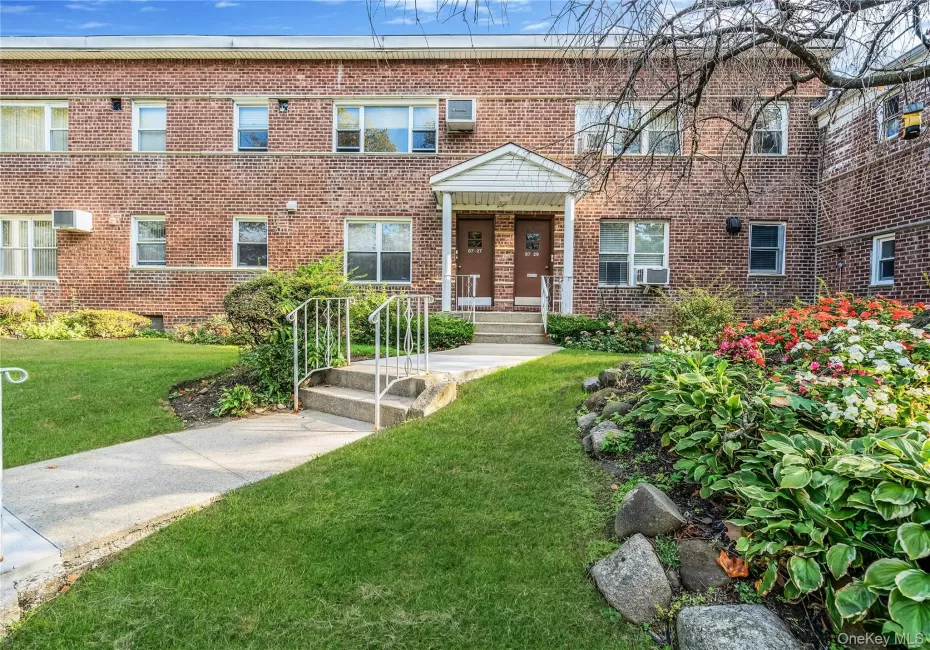 Brick exterior building with a covered entryway, concrete steps, white metal railings, and landscaped grounds featuring mature greenery Brick exterior building with a covered entryway, concrete steps, white metal railings, and landscaped grounds featuring mature greenery