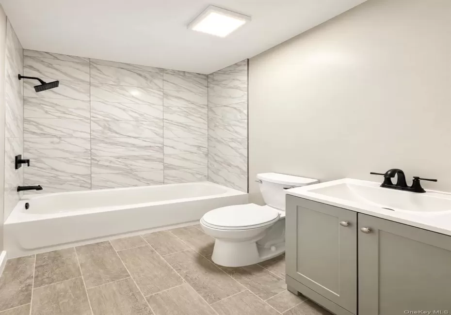 Full bathroom featuring large-format wall tiling in the shower surround, wood-finish flooring, a grey vanity with a white countertop, and matte black plumbing fixtures Full bathroom featuring large-format wall tiling in the shower surround, wood-finish flooring, a grey vanity with a white countertop, and matte black plumbing fixtures