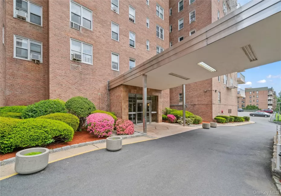 Brick building exterior featuring a covered entryway, decorative landscaping with shrubs and blooming flowers, and an asphalt driveway Brick building exterior featuring a covered entryway, decorative landscaping with shrubs and blooming flowers, and an asphalt driveway