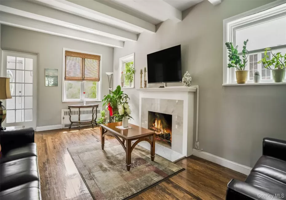 Spacious living area featuring exposed ceiling beams, wood-finish flooring, and a fireplace with a stone surround and white mantel Spacious living area featuring exposed ceiling beams, wood-finish flooring, and a fireplace with a stone surround and white mantel