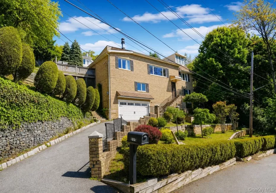Brick façade residence featuring an integrated garage, a terraced front yard, established hedges, and a paved driveway with stone retaining walls Brick façade residence featuring an integrated garage, a terraced front yard, established hedges, and a paved driveway with stone retaining walls