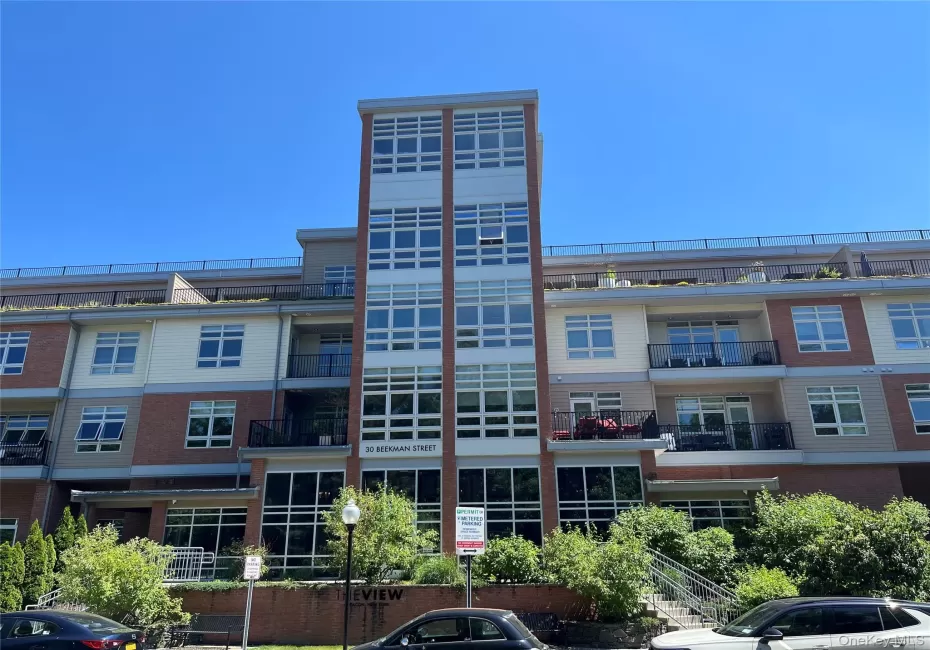 Multi-story residential building featuring a brick and siding exterior, expansive window panels, private balconies with black railings, and a prominent central glass-enclosed stairwell Multi-story residential building featuring a brick and siding exterior, expansive window panels, private balconies with black railings, and a prominent central glass-enclosed stairwell