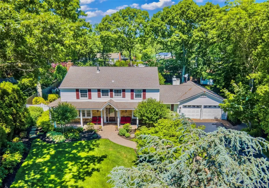 Traditional colonial two-story residence featuring light-colored siding, a covered entry porch, a multi-car garage, mature landscaping, and a paver walkway Traditional colonial two-story residence featuring light-colored siding, a covered entry porch, a multi-car garage, mature landscaping, and a paver walkway