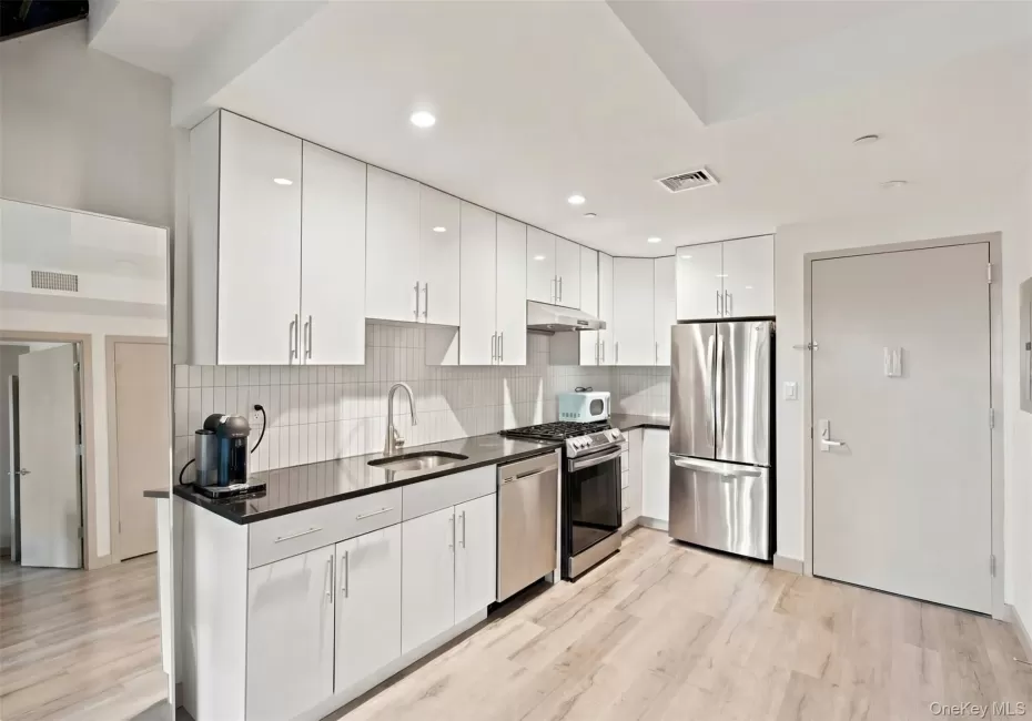 Bright kitchen featuring glossy white cabinetry, dark countertops, white subway tile backsplash, stainless steel appliances, and light wood-finish flooring Bright kitchen featuring glossy white cabinetry, dark countertops, white subway tile backsplash, stainless steel appliances, and light wood-finish flooring