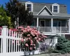 Gambrel roofline property featuring a full-width front porch with white columns, light grey shingle siding, and two dormer windows. Perennial rose bush in bloom. Gambrel roofline property featuring a full-width front porch with white columns, light grey shingle siding, and two dormer windows. Perennial rose bush in bloom.