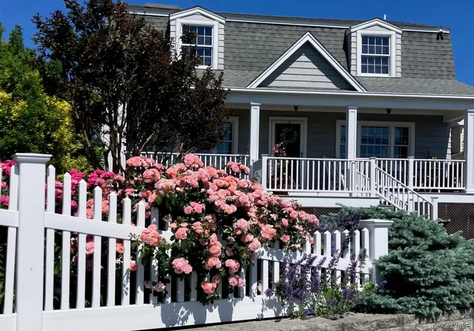 Gambrel roofline property featuring a full-width front porch with white columns, light grey shingle siding, and two dormer windows. Perennial rose bush in bloom. Gambrel roofline property featuring a full-width front porch with white columns, light grey shingle siding, and two dormer windows. Perennial rose bush in bloom.