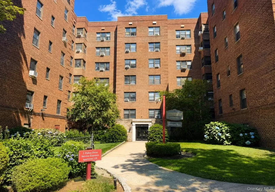 Brick facade building featuring multiple window styles, a central entryway with an arched stone surround, a paved walkway, manicured lawn areas, and established shrubbery Brick facade building featuring multiple window styles, a central entryway with an arched stone surround, a paved walkway, manicured lawn areas, and established shrubbery