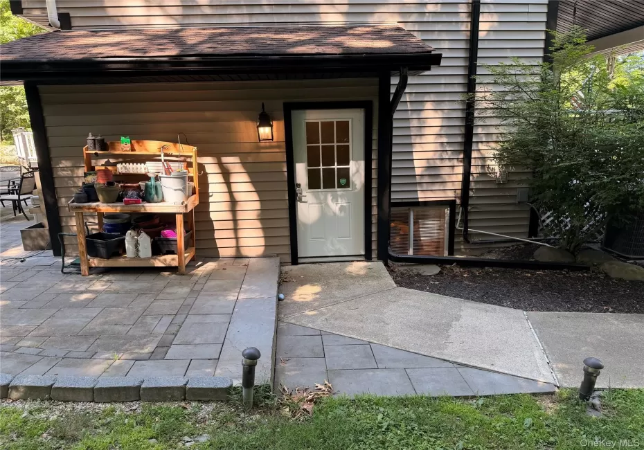 Outdoor paver patio with a concrete walkway, exterior wall light fixture, and a white door with window panes Outdoor paver patio with a concrete walkway, exterior wall light fixture, and a white door with window panes