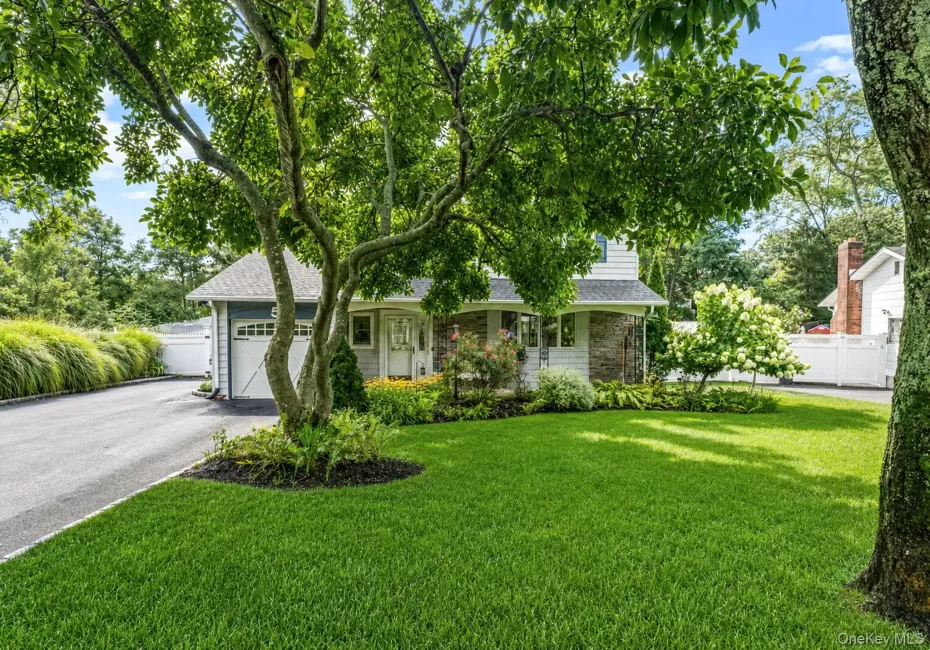 Manicured front lawn with mature shade trees, paved driveway, stone facade accents, white picket fencing, and a garage with a carriage-style door Manicured front lawn with mature shade trees, paved driveway, stone facade accents, white picket fencing, and a garage with a carriage-style door