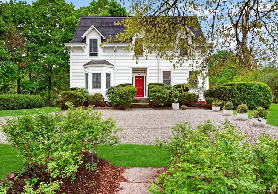 Exterior of colonial residence with red door and mansard roof. Gravel driveway with manicured gardens surrounding. Exterior of colonial residence with red door and mansard roof. Gravel driveway with manicured gardens surrounding.