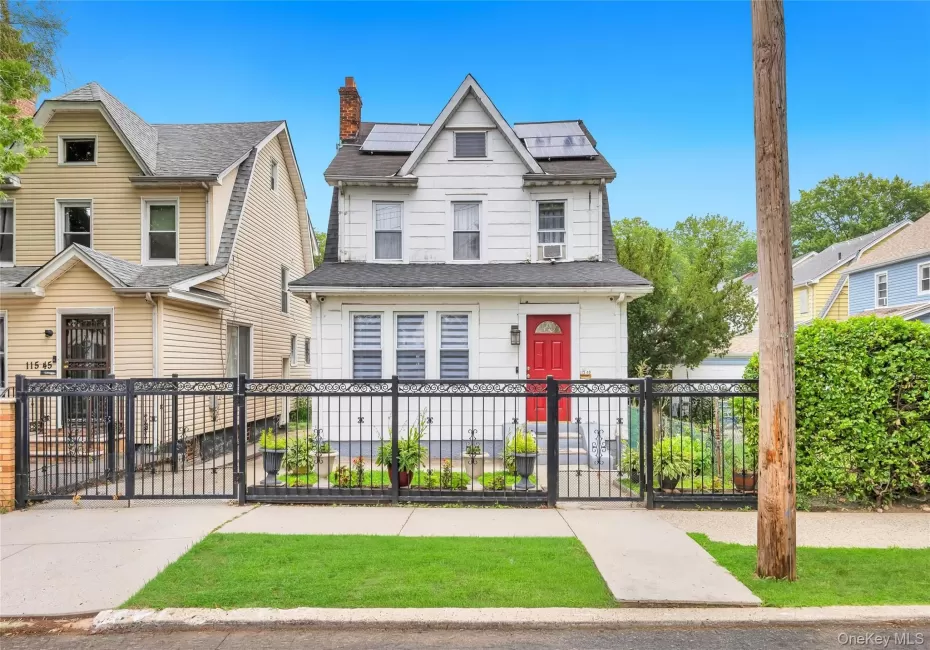 White two-story residence featuring a prominent red entry door, an ornate black metal fence, and solar panels White two-story residence featuring a prominent red entry door, an ornate black metal fence, and solar panels