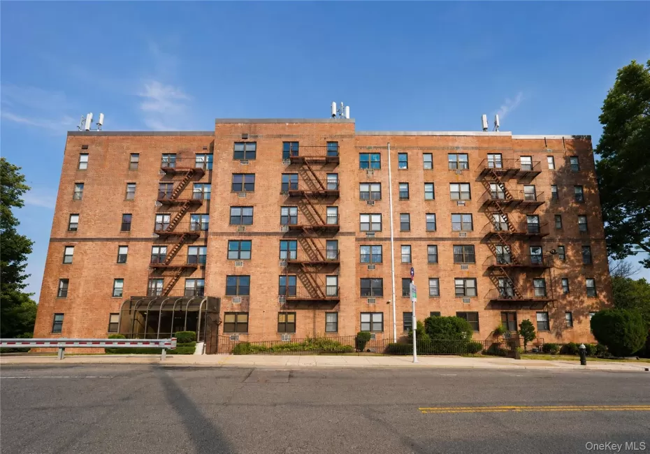 Brick multi-story building featuring multiple windows, exterior fire escapes, a glass-enclosed entrance, and surrounding shrubbery Brick multi-story building featuring multiple windows, exterior fire escapes, a glass-enclosed entrance, and surrounding shrubbery