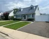 Classic Cape Cod home featuring a freshly sealed asphalt driveway, manicured lawn with defined landscaping borders, large picture windows with dark trim, dormers with vinyl siding, and an attached garage with white paneled doors Classic Cape Cod home featuring a freshly sealed asphalt driveway, manicured lawn with defined landscaping borders, large picture windows with dark trim, dormers with vinyl siding, and an attached garage with white paneled doors