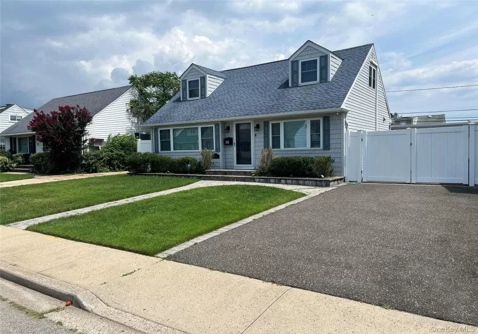 Classic Cape Cod home featuring a freshly sealed asphalt driveway, manicured lawn with defined landscaping borders, large picture windows with dark trim, dormers with vinyl siding, and an attached garage with white paneled doors Classic Cape Cod home featuring a freshly sealed asphalt driveway, manicured lawn with defined landscaping borders, large picture windows with dark trim, dormers with vinyl siding, and an attached garage with white paneled doors